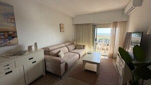 Una sala de estar con sofá, mesa de centro y televisor. Hay un balcón con vista a la playa.