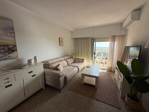 Ein Wohnzimmer mit einer Couch, einem Couchtisch und einem Fernseher. Ein Balkon bietet Blick auf den Strand.