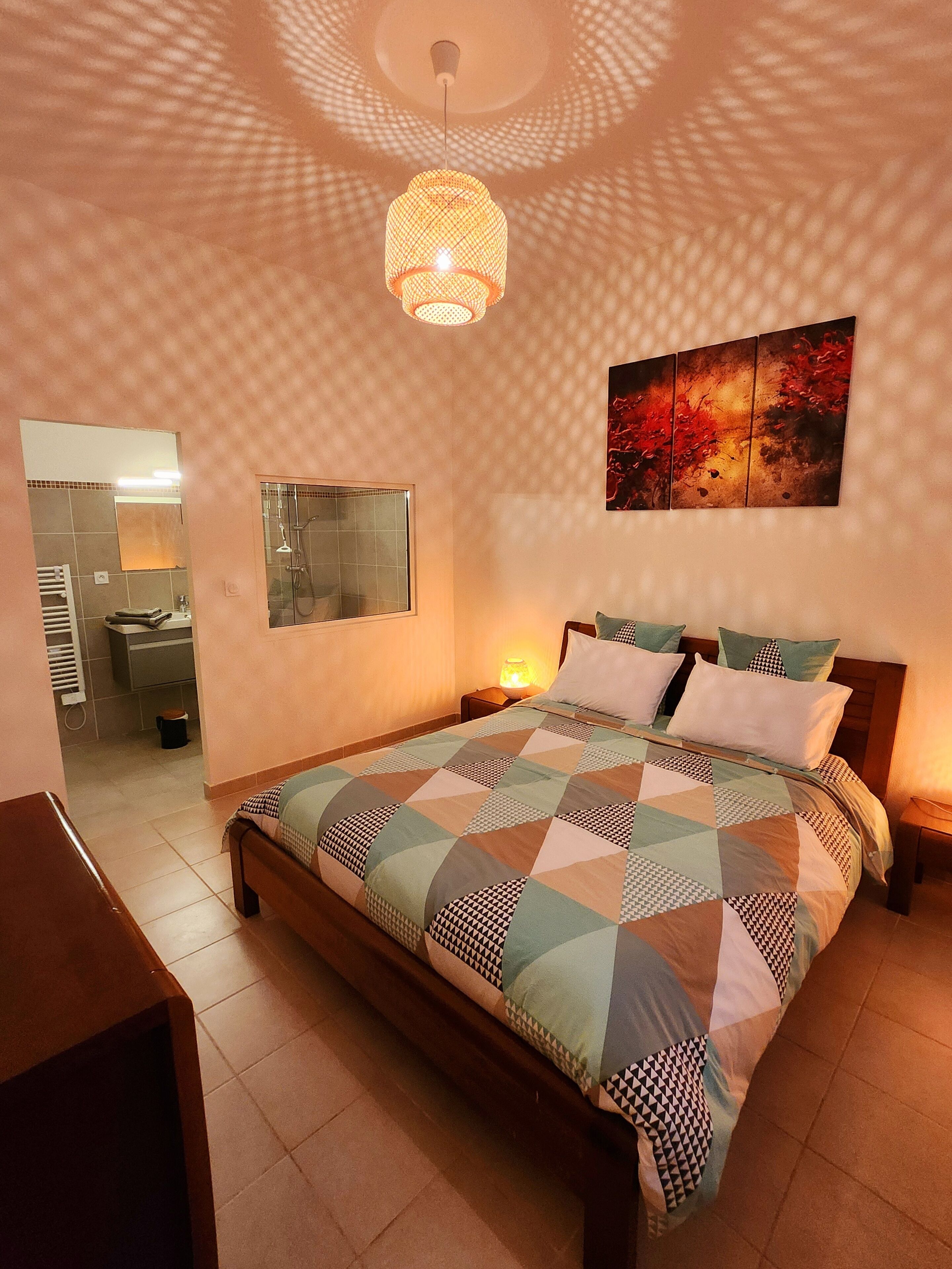 Ein Schlafzimmer mit einem Bett, einem Nachttisch, einem Wandbild und einem durch eine offene Tür einsehbaren Badezimmer.