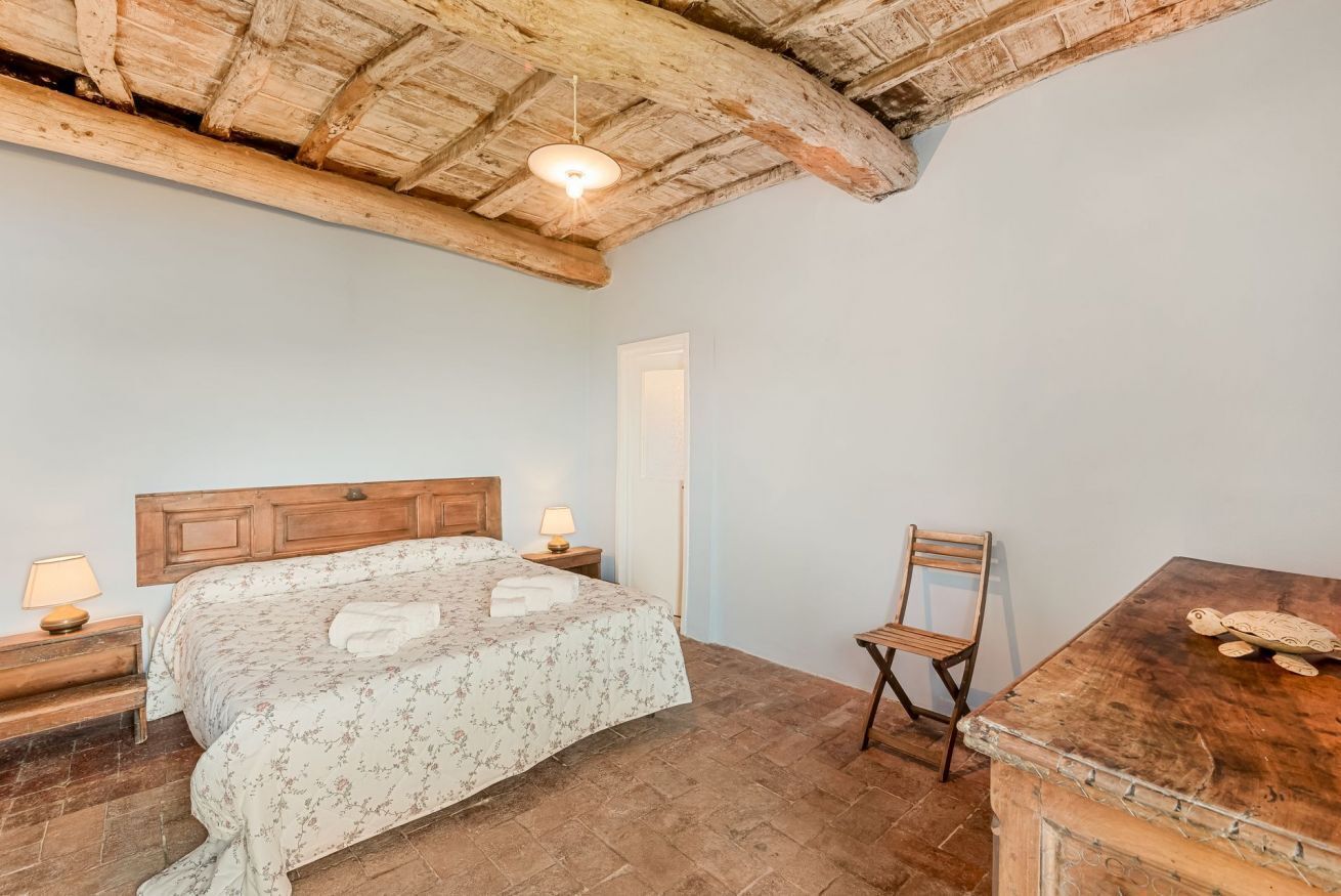 Ein Schlafzimmer mit einem Holzkopfende, einem Bett mit floralem Bettbezug, einem Holzstuhl, einem Holztisch und einer rustikalen Decke.