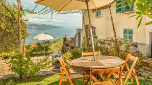 Ein Holztisch mit Stühlen steht unter einem großen Sonnenschirm, mit Blick auf das Meer und einem Steinhaus im Hintergrund.