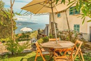 A wooden table and chairs under a large umbrella, with a view of the sea and a stone building in the background. - [ilCeppo] 5Terre-Seaview-Parking (La Spezia)
