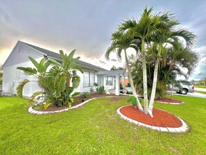 A residential property with a white exterior, a covered patio, and a lawn with palm trees. - Cute House in East Englewood (Port Charlotte Thorman Rd)