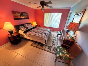 A bedroom with a bed, two bedside lamps, a ceiling fan, a window with blinds, a chair, and a nightstand. - Cute House in East Englewood (Port Charlotte Thorman Rd)