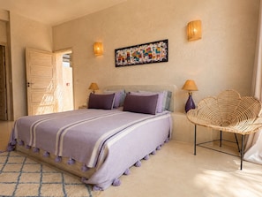 Un dormitorio con una cama, dos lámparas de pared, una obra de arte colorida y una silla de mimbre.