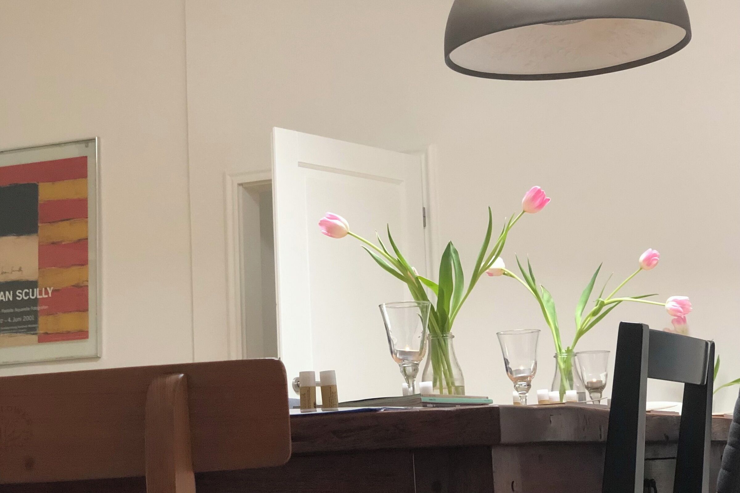 A dining area with a table set for two, a mirror, and a painting on the wall.