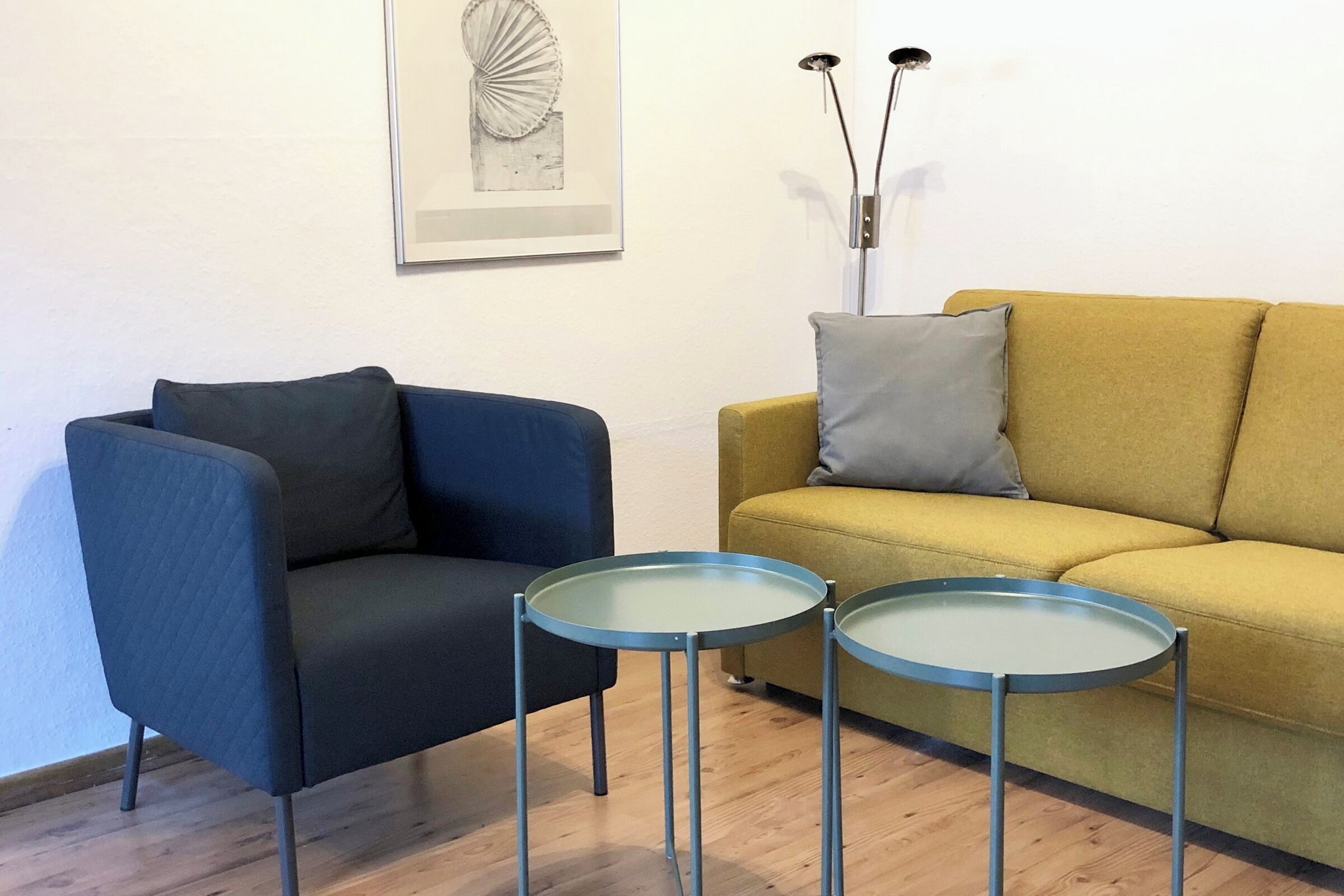 Ein Wohnzimmer mit blauem Sessel, gelbem Sofa und zwei kleinen runden Tischen.