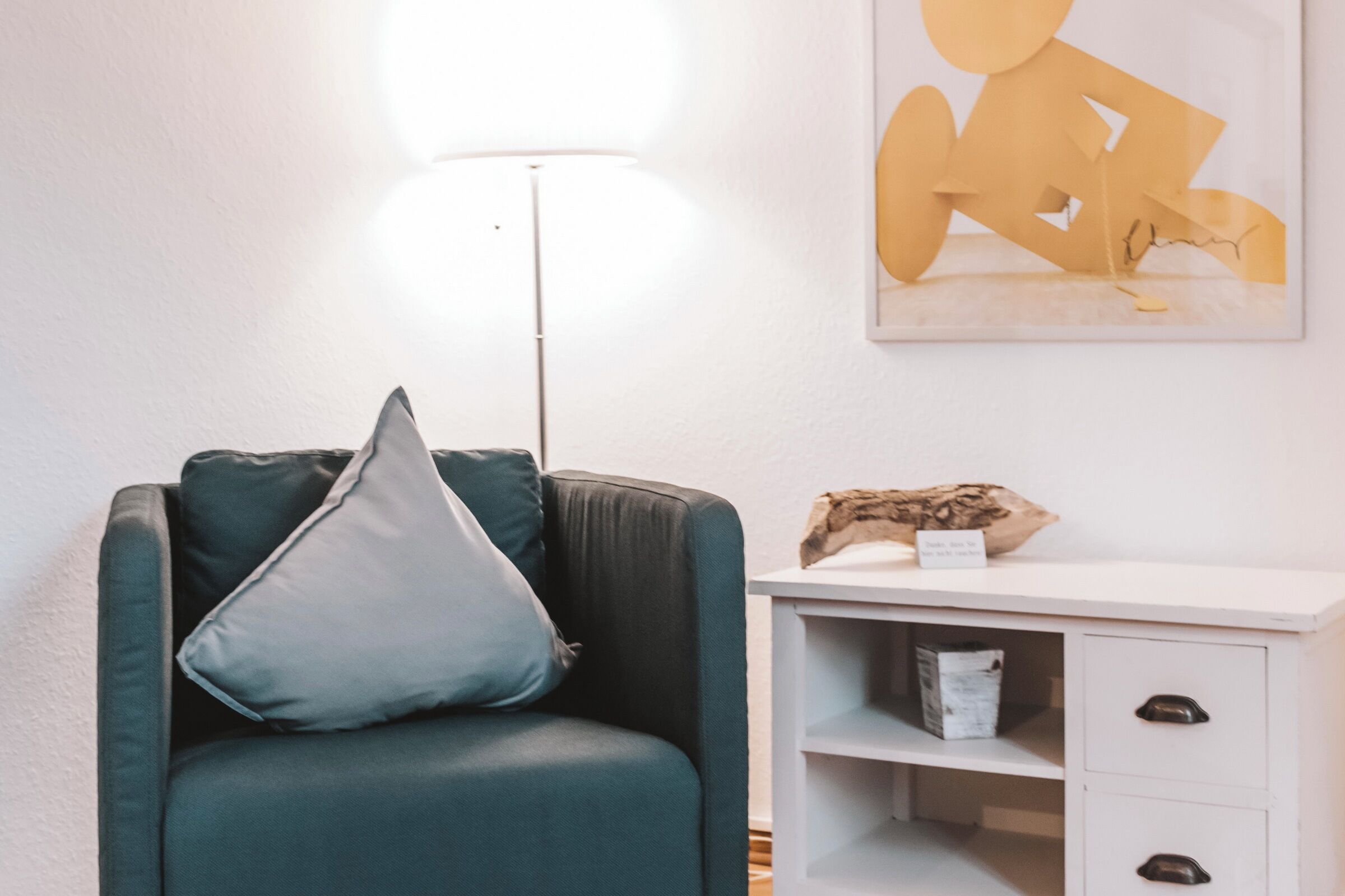 Una habitación con un sillón verde oscuro, una mesita auxiliar blanca con un objeto decorativo, y un cuadro abstracto colgado en la pared.