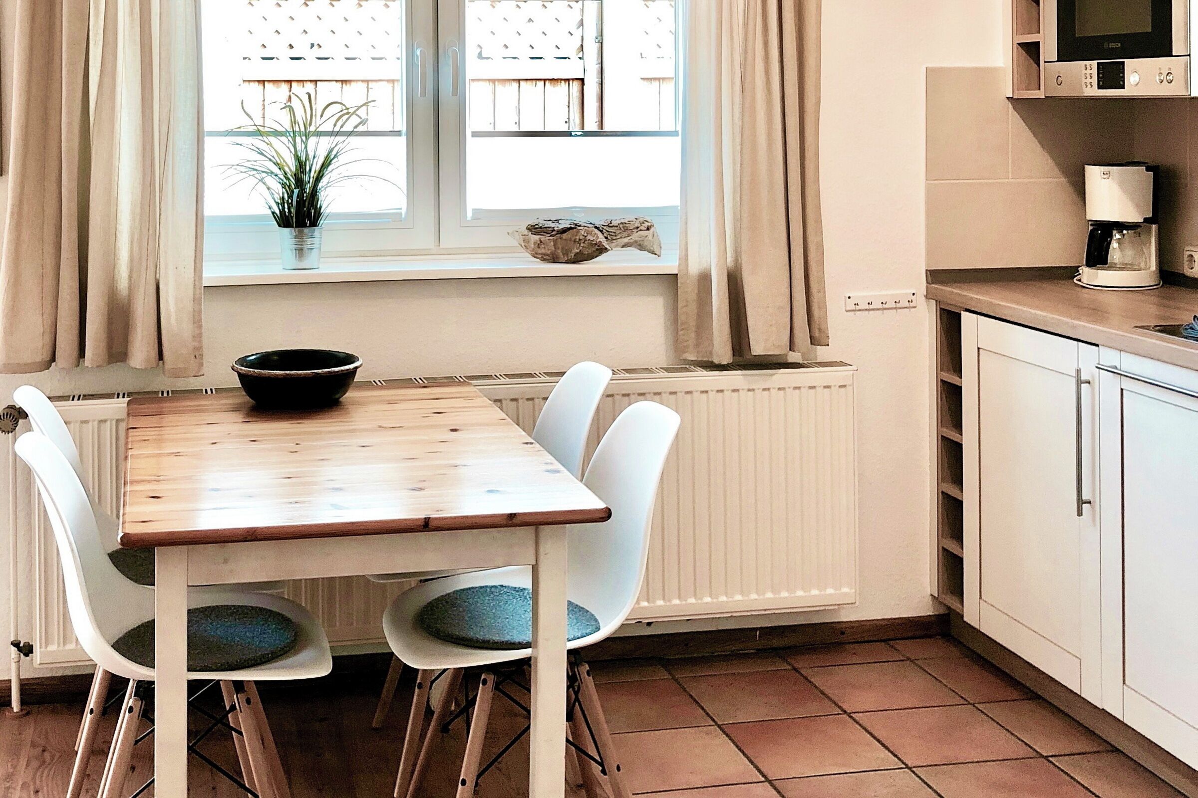 Eine Küche mit einem Holztisch, weißen Stühlen, einem Fenster mit Vorhängen, einer Topfpflanze und einer Kaffeemaschine.