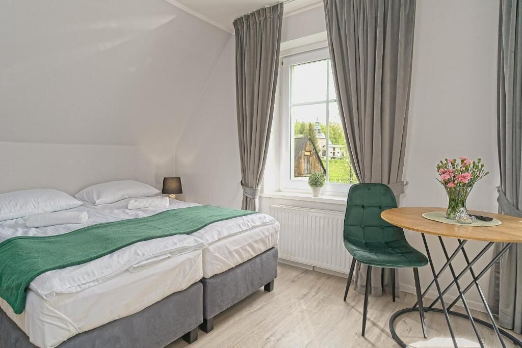 A bedroom with a bed, a bedside table, a chair, a window with curtains, and a view of greenery outside.