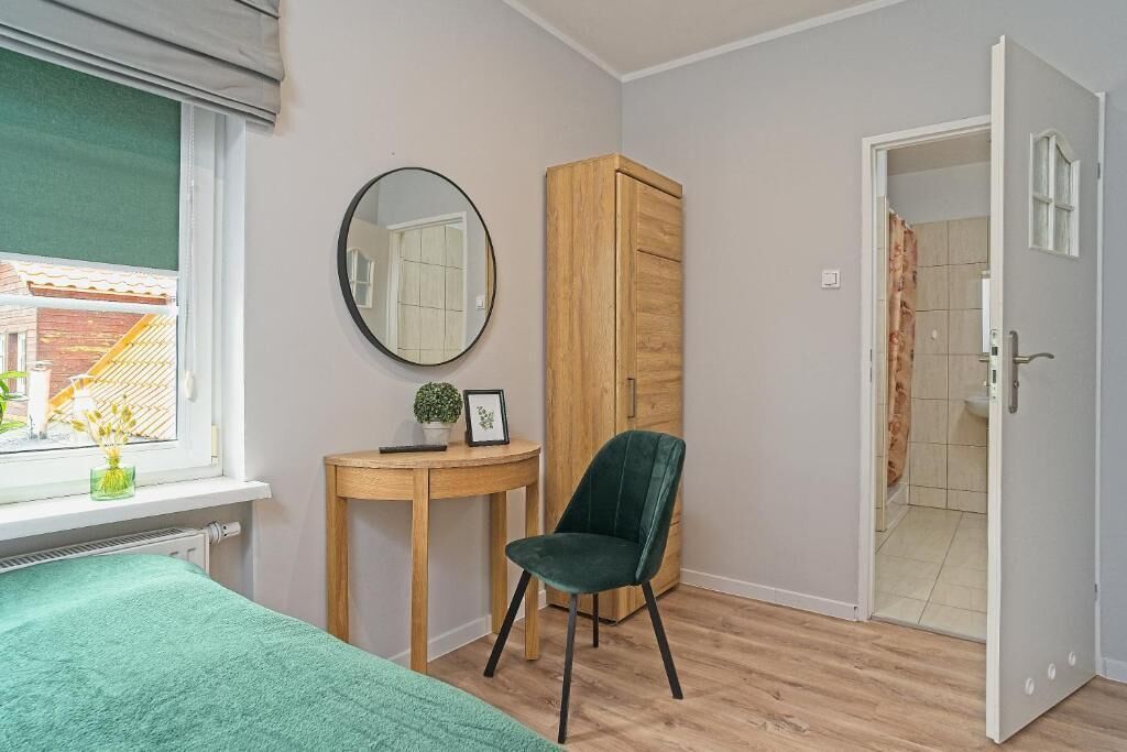 A bedroom with a bed, a wooden wardrobe, a round mirror, a small table with a plant, and a chair.