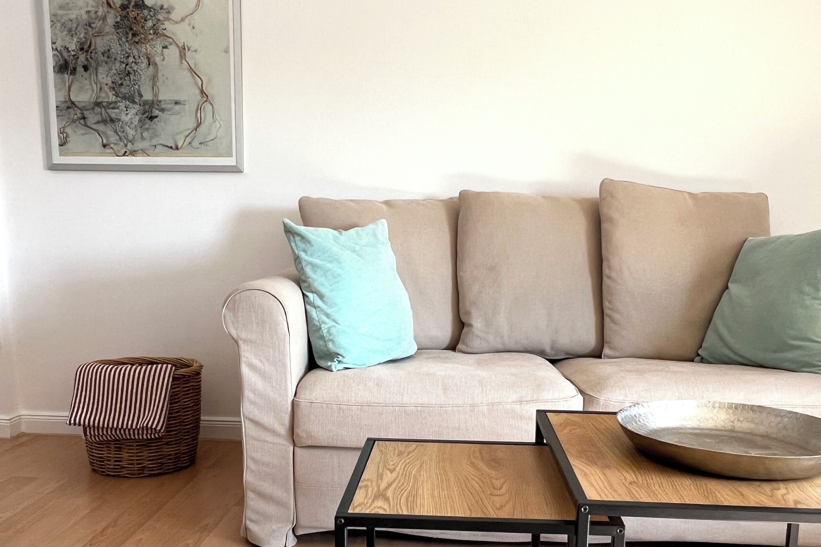 Un salon avec un canapé beige, un panier à rayures, un tableau abstrait encadré et une table basse en bois sur laquelle repose un bol en métal.