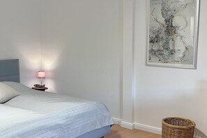 Et soveværelse med en seng, et natbord med en lampe, en fletkurv og et abstrakt kunstværk i ramme på væggen.