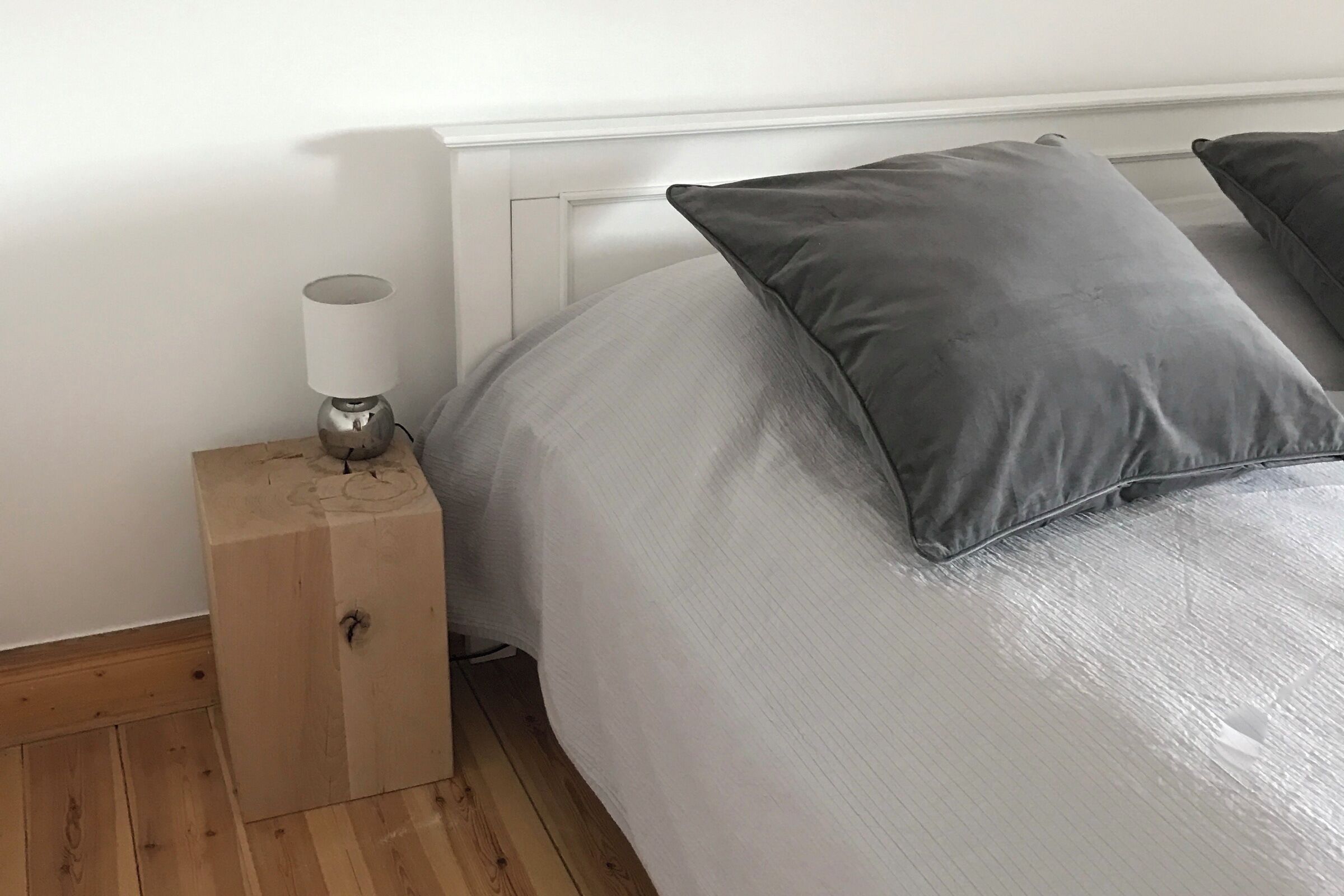 Una cama bien hecha con ropa de cama gris, una lámpara de noche y una mesita de noche de madera.