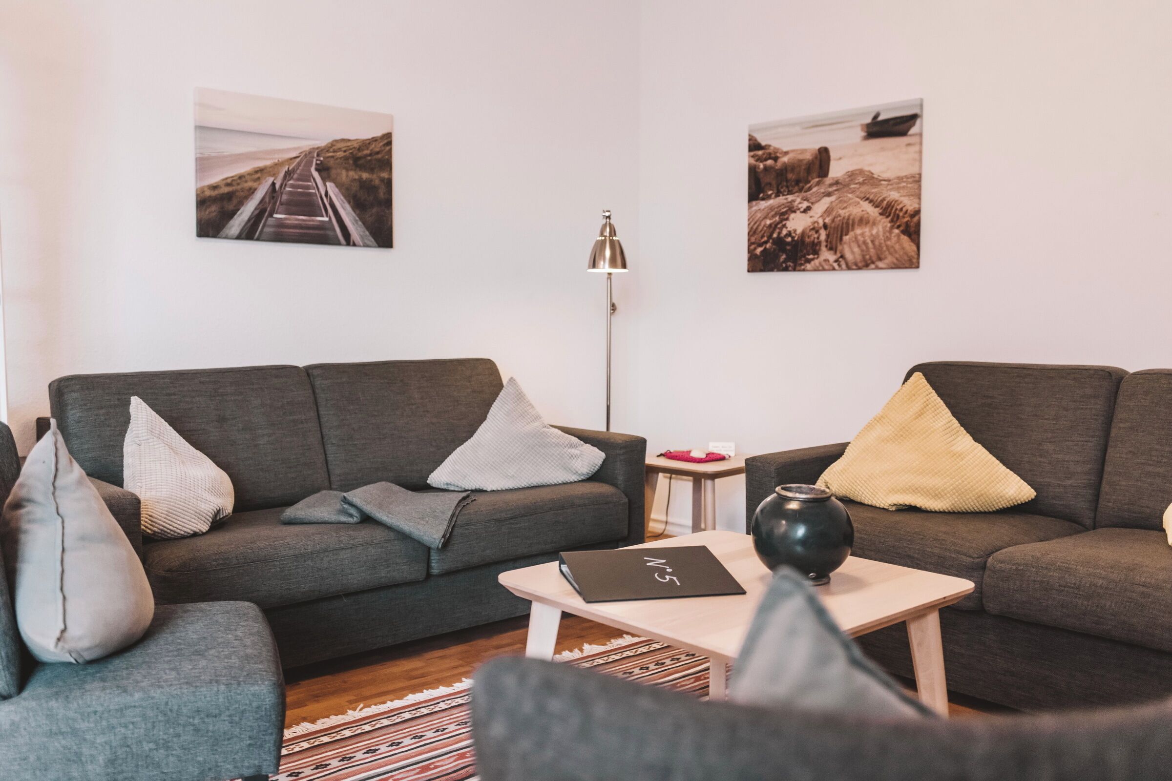 A modern living room with two grey sofas, a small coffee table, and a floor lamp. There are two framed pictures on the wall.