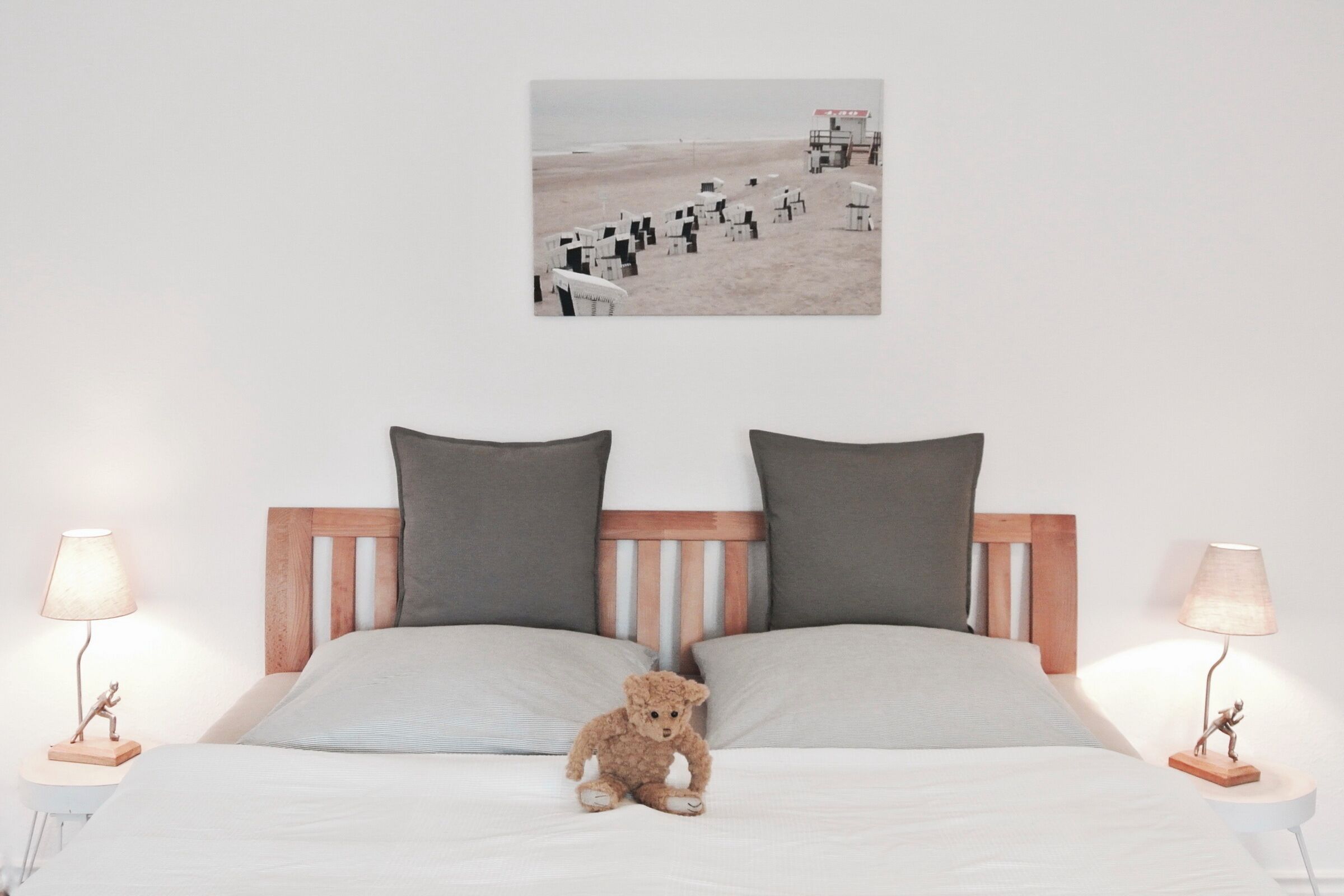 Un dormitorio con cabecera de madera, dos almohadas, un pequeño osito de peluche y una pintura de una playa en la pared.