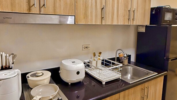A kitchen with wooden cabinets, a black countertop, a sink, a stove, and a refrigerator. - Shore 2 Cozy Staycation, Pool View, Near MOA and NAIA Airport w/ Netflix (Pasay City)