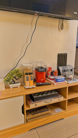 A wooden desk with a computer monitor, a red appliance, a BINGO set, a Rubik's Cube, and a wooden word game. - Shore 2 Cozy Staycation, Pool View, Near MOA and NAIA Airport w/ Netflix (Pasay City)