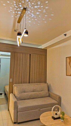 A modern living room with a sofa, a wooden cabinet, and a ceiling light fixture. - Shore 2 Cozy Staycation, Pool View, Near MOA and NAIA Airport w/ Netflix (Pasay City)