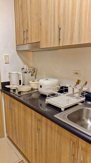 A kitchen with wooden cabinets, a stainless steel sink, a coffee maker, a rice cooker, and a white appliance on the countertop. - Shore 2 Cozy Staycation, Pool View, Near MOA and NAIA Airport w/ Netflix (Pasay City)