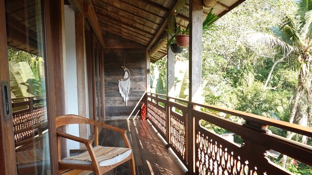 A wooden balcony with a chair, a decorative wall hanging, and a view of lush greenery. - Villa Cempaka Water Portal Ubud (Gianyar)