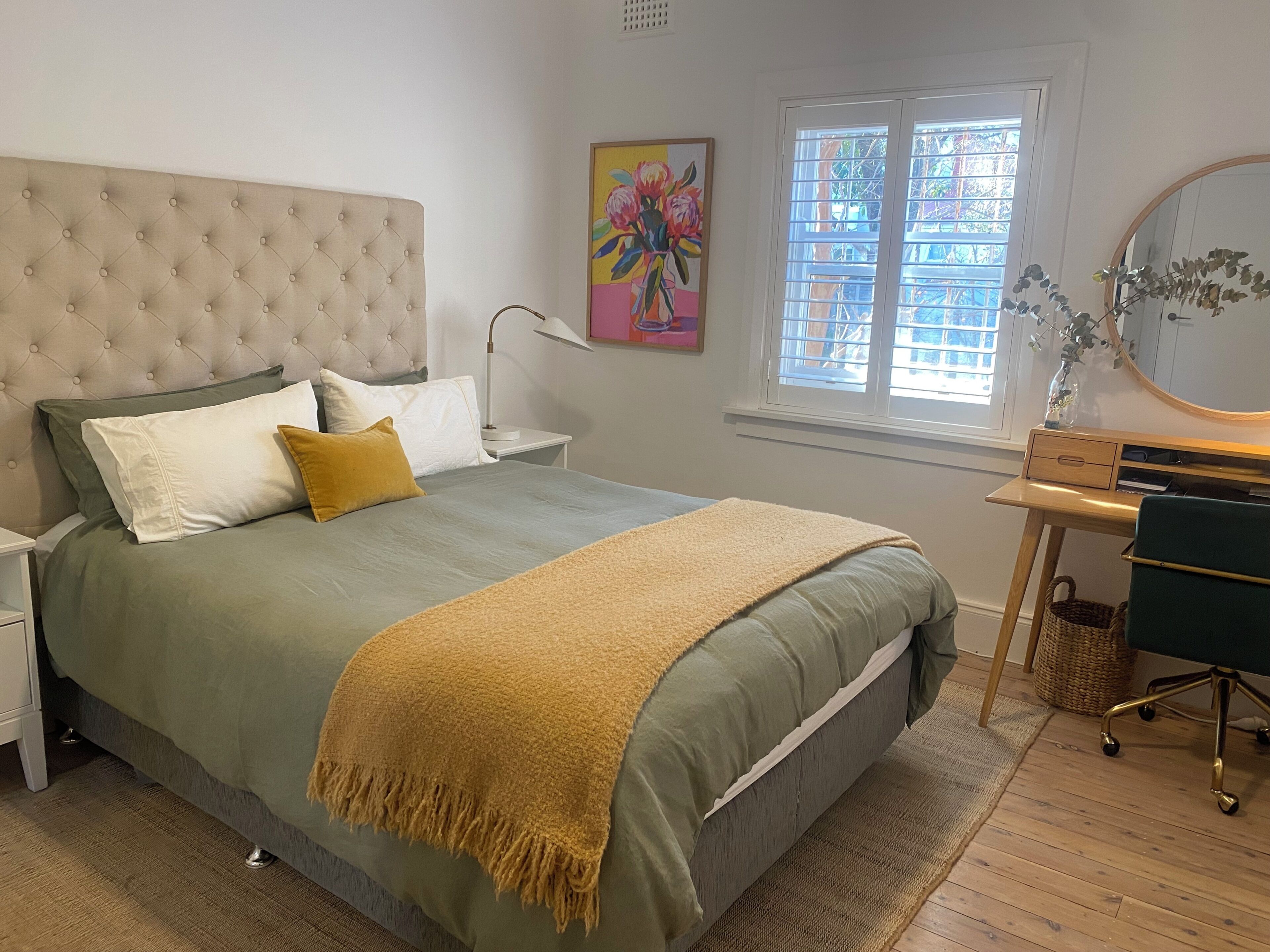 Une chambre à coucher avec une tête de lit capitonnée, un lit avec une literie verte, un plaid jaune, un bureau avec une chaise et une fenêtre avec des stores.