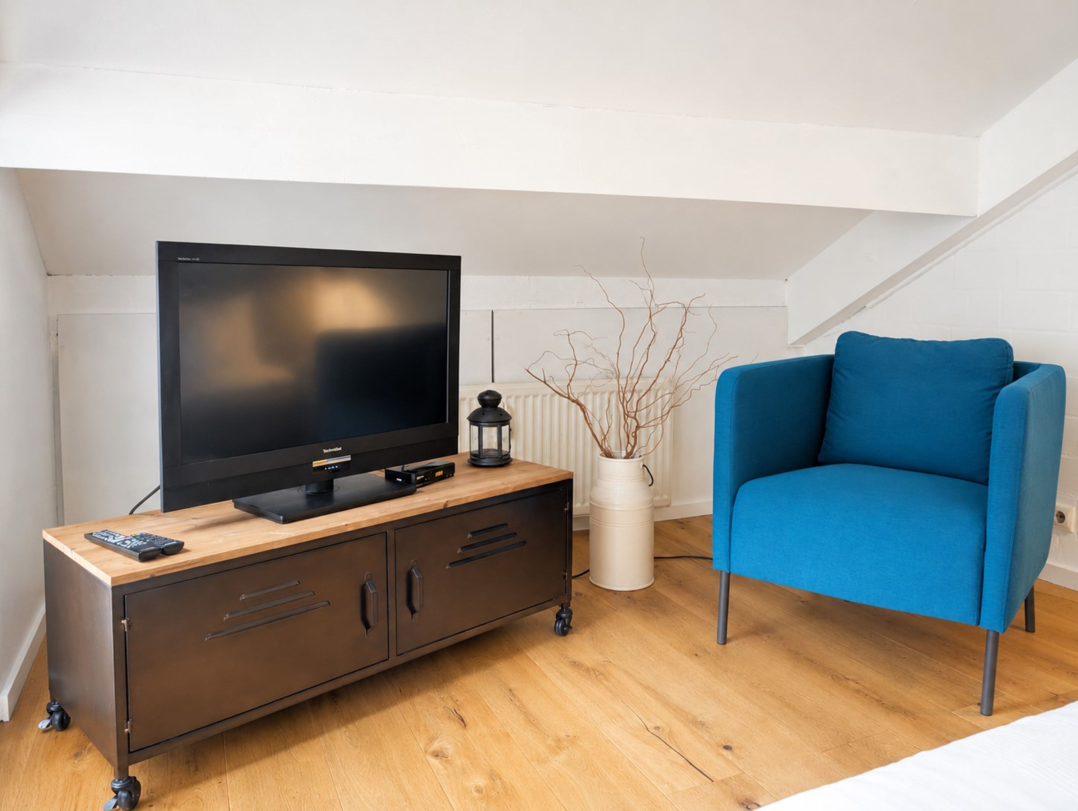 Ein modernes Wohnzimmer mit einem Flachbildfernseher auf einer Holzkommode, einem blauen Sessel und einer weißen Vase mit Zweigen.