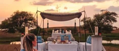 Disposición para comer al aire libre con una mesa y sillas, una persona sentada, y una mesa con comida y bebidas.