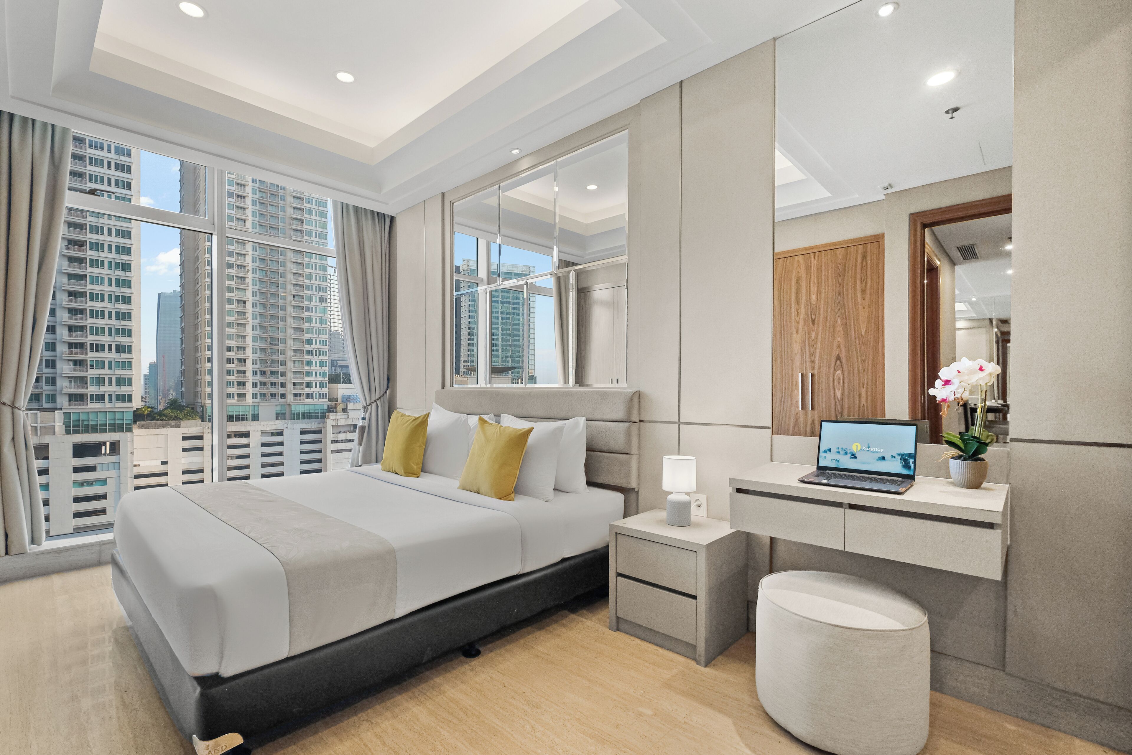 A modern hotel room with a large bed, a desk, a laptop, and a view of high-rise buildings.