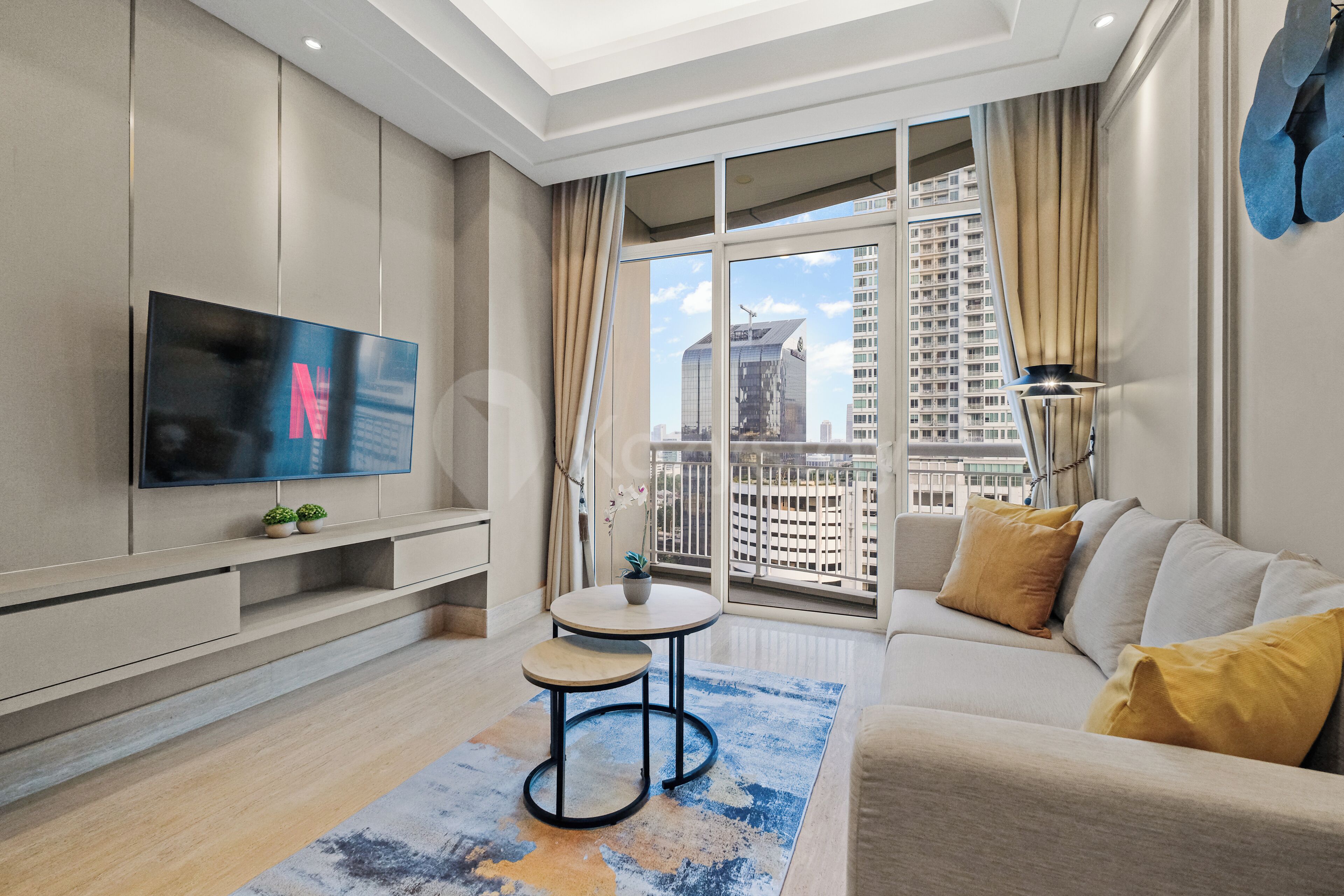 A modern living room with a flat-screen TV, a sofa, a coffee table, and a view of a cityscape through large windows.