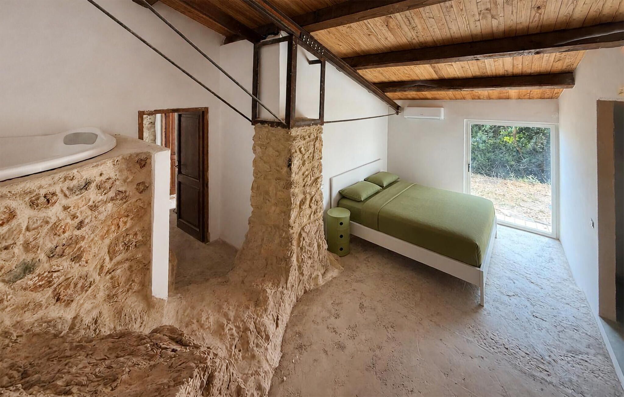 Ein Schlafzimmer mit Stehwand, einem Bett mit grüner Bettwäsche, einem Fenster und einer Holzbalkendecke.