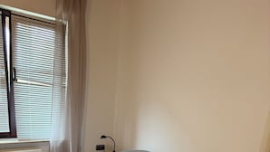 Ein Einzelbett mit einem gemusterten Überwurf und Kissen, ein Nachttisch mit Lampe sowie ein Fenster mit Jalousien und Vorhängen.