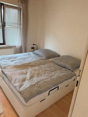 Ein Einzelbett mit einem gemusterten Überwurf und Kissen, ein Nachttisch mit Lampe sowie ein Fenster mit Jalousien und Vorhängen.