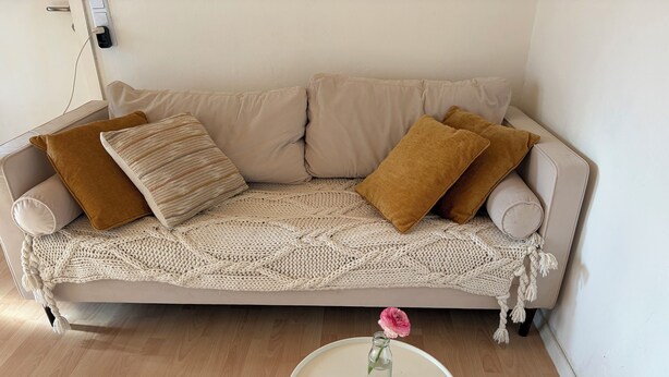 Ein beiges Sofa mit einer gestrickten Decke, zwei braunen Kissen und einem kleinen weißen Tisch mit einer rosa Blumenvase.