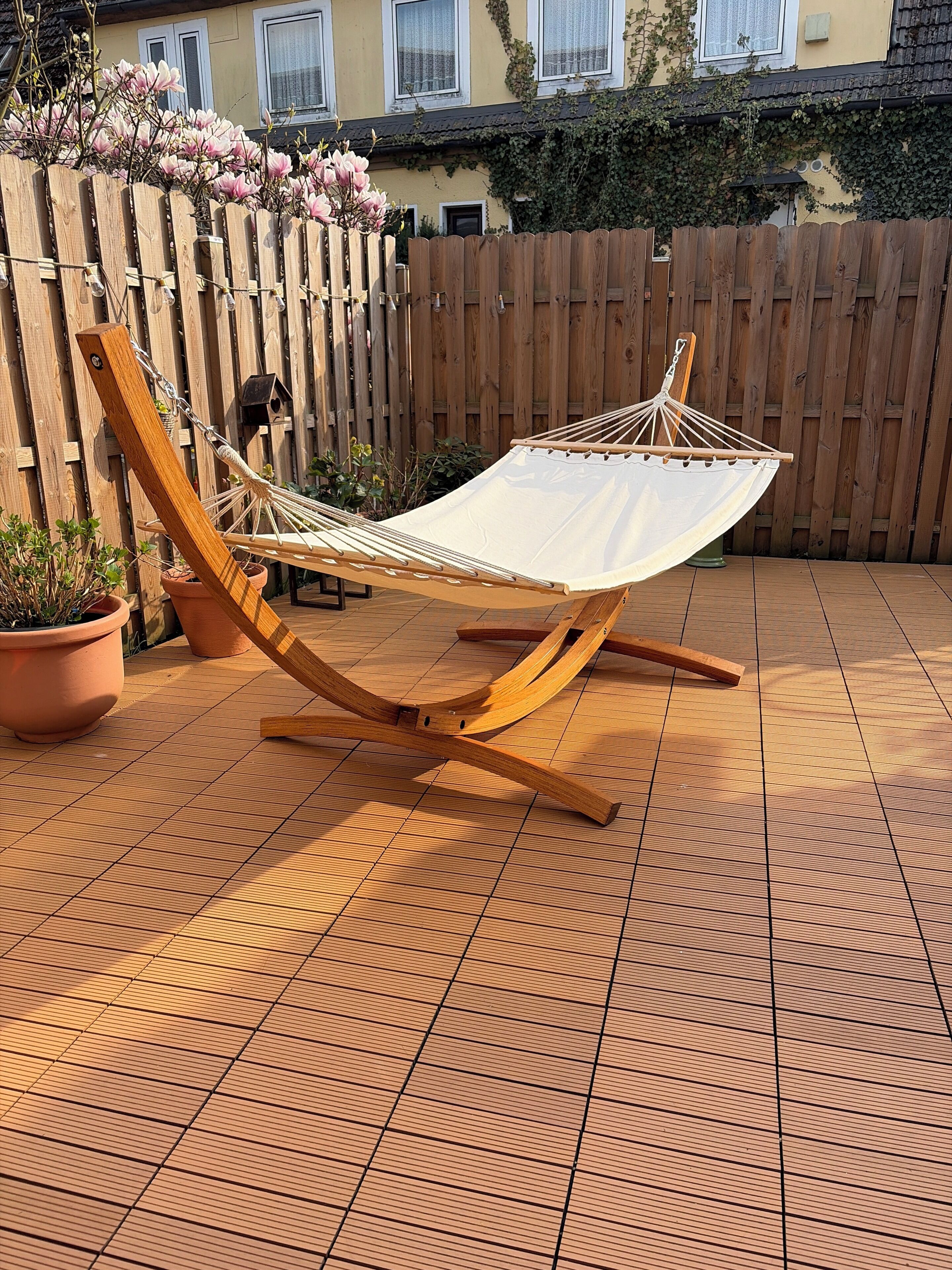 Una hamaca con tela blanca, estructura de madera y cadenas de metal, ubicada en un patio empedrado con plantas en macetas y una valla de madera al fondo.