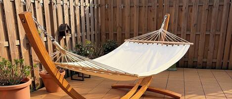 A hammock with a white fabric, wooden frame, and metal chains, set on a tiled patio with potted plants and a wooden fence in the background.