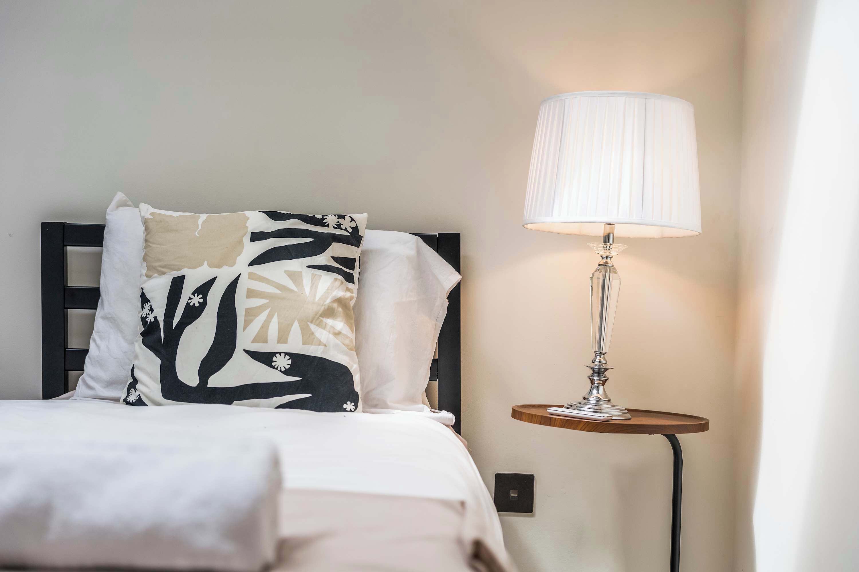 Een bed met een kussen met een zwart-wit patroon, een wit kussen en een witte spreideken. Er staat een lamp met een witte lampenkap op een houten nachtkastje naast het bed.