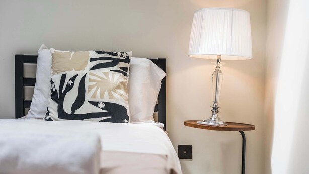 A bed with a white and black patterned pillow, a white pillow, and a white bedspread. There is a lamp with a white shade on a wooden side table beside the bed. - Modern Flat In Jewellery Quarter, Free Parking (Birmingham)