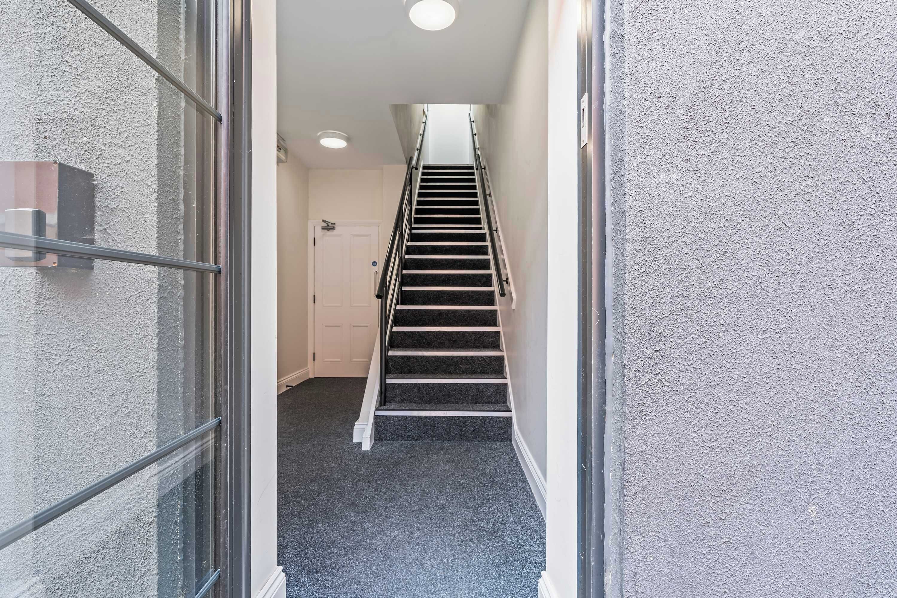A narrow hallway leading to a staircase with a handrail, a door at the end, and a textured wall on one side.
