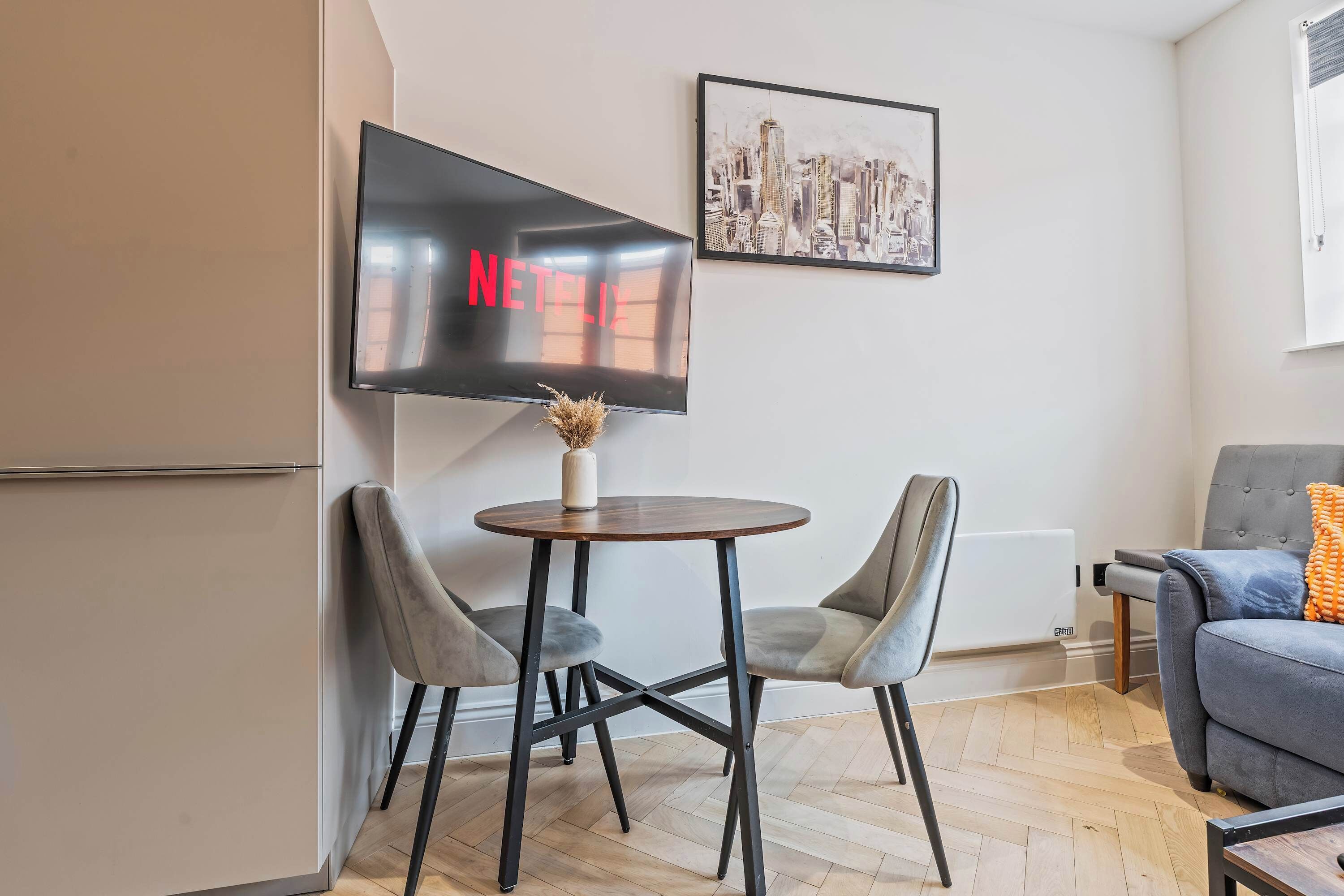 A modern living room with a flat-screen TV mounted on the wall, a round wooden table with two chairs, a grey sofa, and a framed cityscape picture.