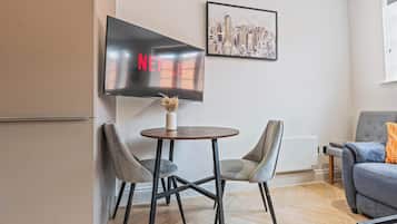 A modern living room with a flat-screen TV mounted on the wall, a round wooden table with two chairs, a grey sofa, and a framed cityscape picture.