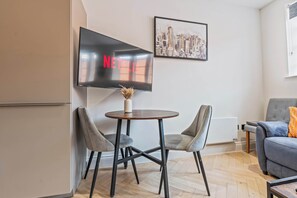 A modern living room with a flat-screen TV mounted on the wall, a round wooden table with two chairs, a grey sofa, and a framed cityscape picture. - Modern Flat In Jewellery Quarter, Free Parking (Birmingham)