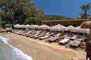 Un resort frente al mar con filas de tumbonas y sombrillas de paja, una playa arenosa y un fondo de árboles y colinas.