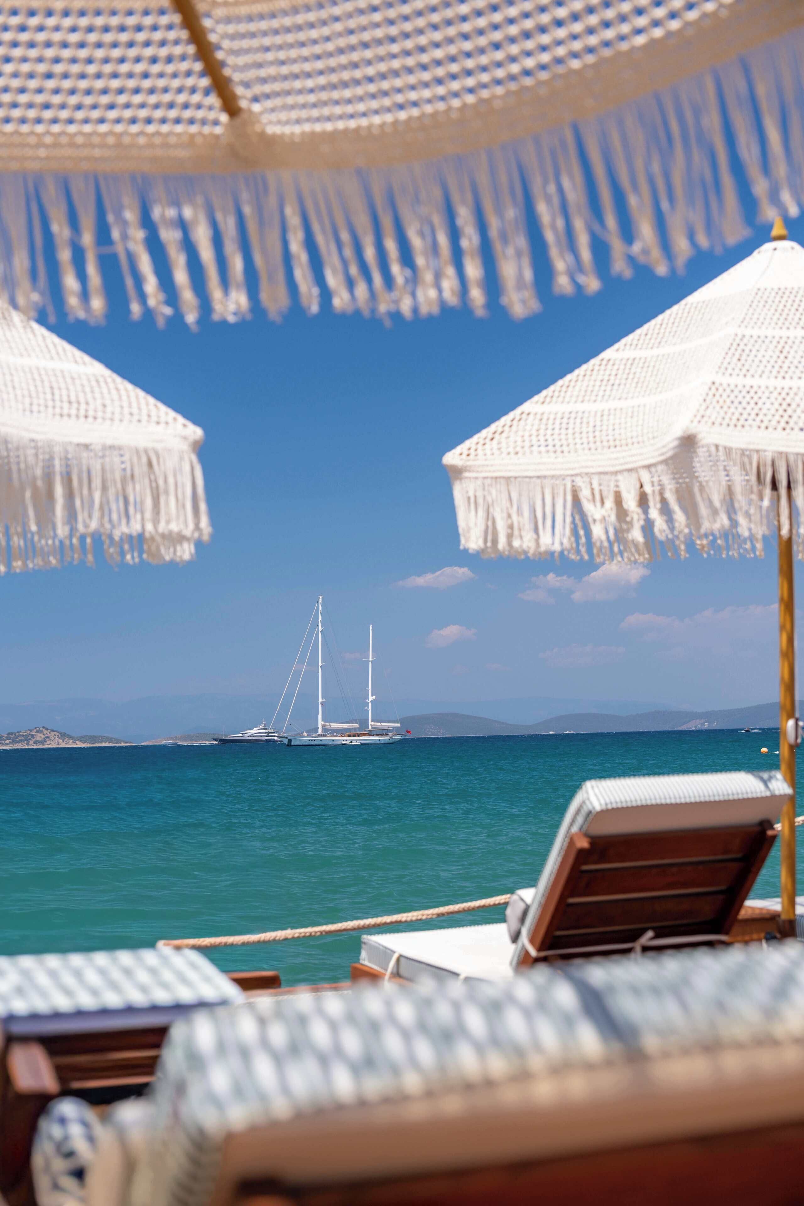 A beachside view with two white umbrellas, a sailboat in the distance, and a clear blue sky.