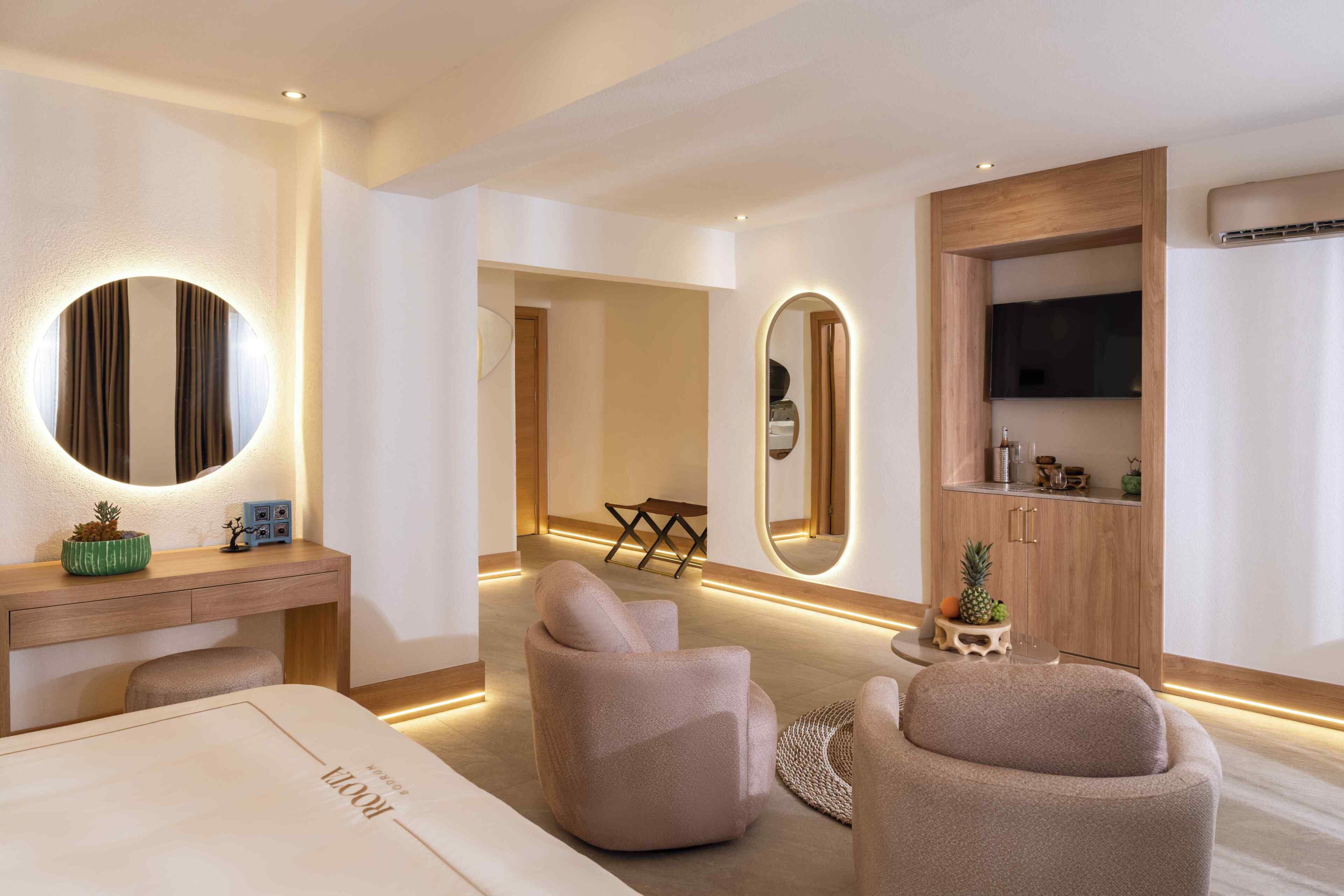 A modern hotel room with a large bed, two armchairs, a wooden vanity with a mirror, and a television on a wooden cabinet.