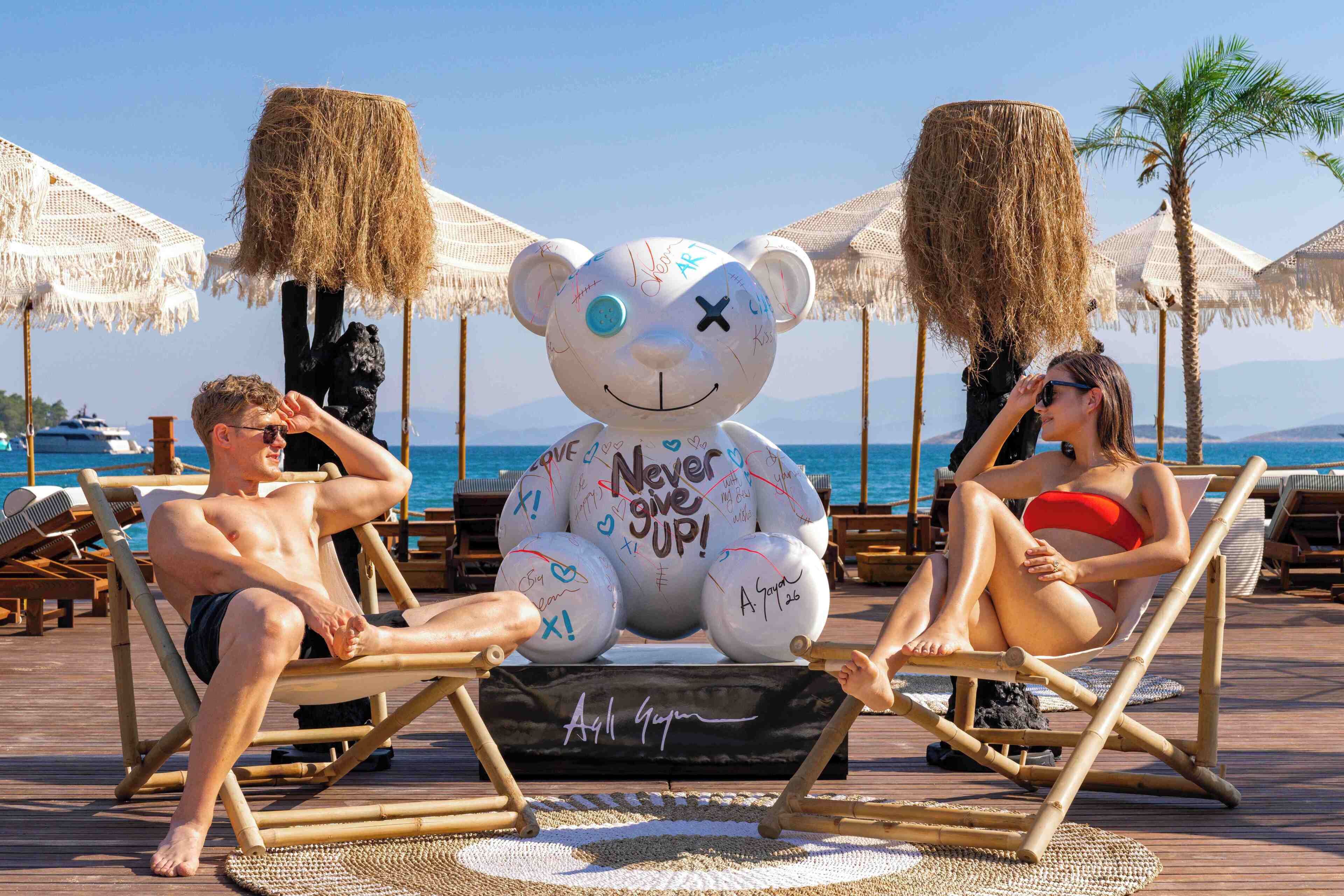 A beach scene with a large white bear sculpture with graffiti, two people sitting on lounge chairs, and a clear view of the sea.