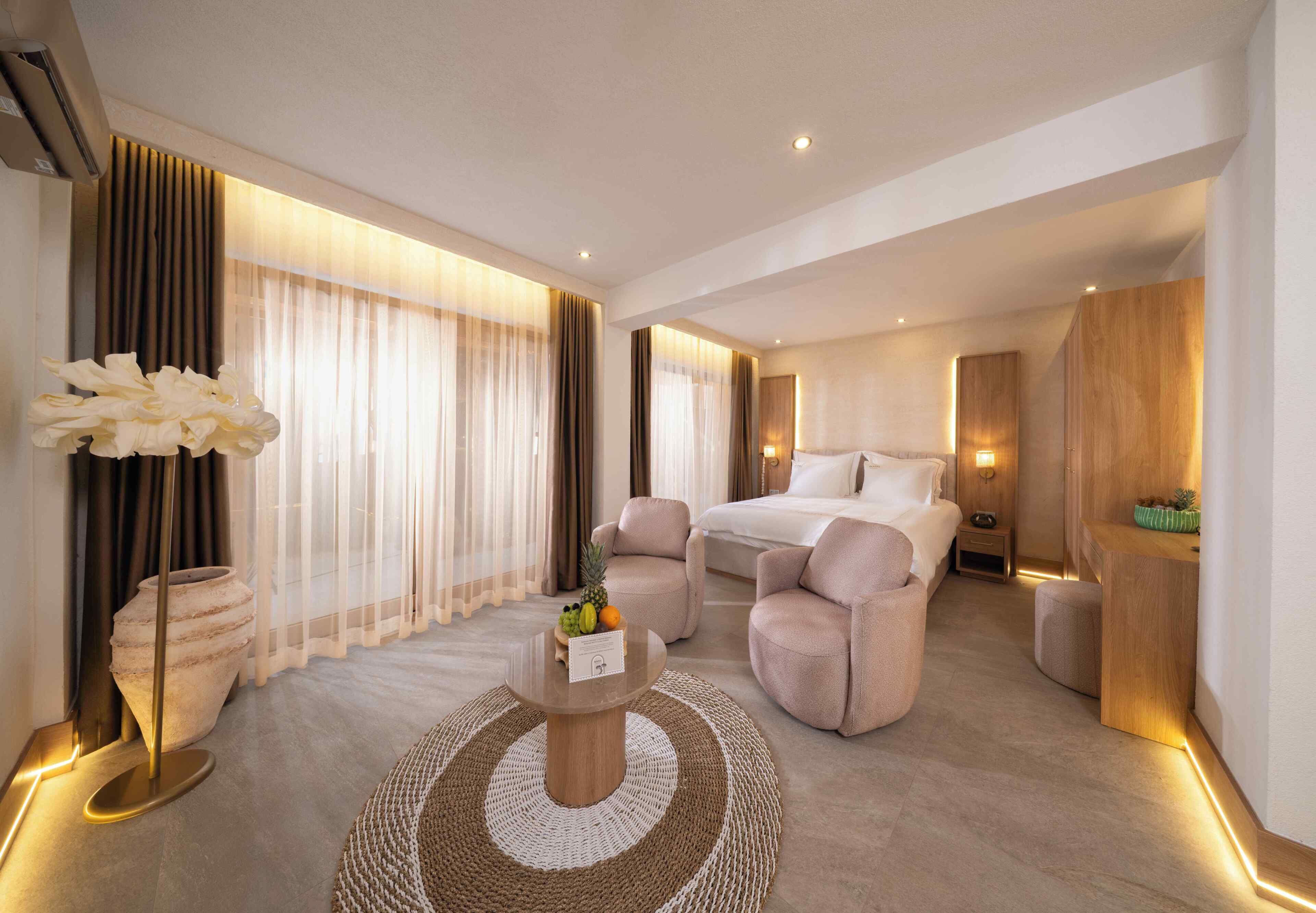 Ein modernes Hotelzimmer mit einem großen Bett, zwei Sesseln, einem Couchtisch mit Obst und Blick auf die Stadt.