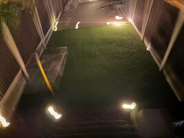 Un sendero estrecho al aire libre con iluminación artificial que conduce a una zona verde, rodeado de muros.