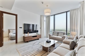 A modern living room with a large flat-screen TV, a sofa set, a coffee table, and a view of the city through large windows. - 2BR Apartment With iconic view+Ample of amenities (Dubai)