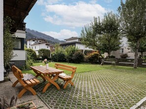 A patio with a wooden table and benches, surrounded by a garden with grass and trees. - Haus Adler 2 by Alpine Host Helpers (Westendorf)